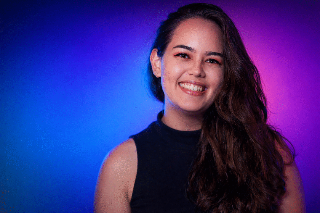 Retrato em close de mulher sorrindo, com cabelo castanho longo de lado, iluminada por luz azul de um lado e roxa do outro, sobre fundo em degradê azul e magenta.